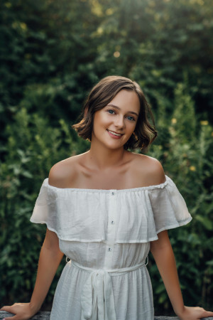 Cassidy, in a white dress, standing in front of a forest and smiling.