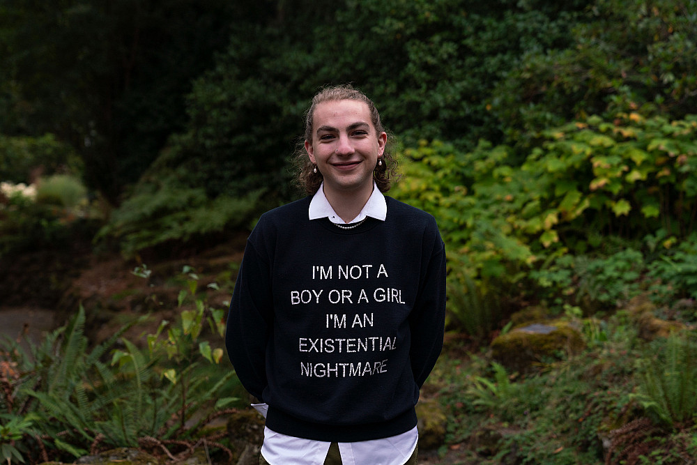 Logan smiling outside. They are wearing a sweater that says I'm not a boy or a girl. I'm an existential nightmare.