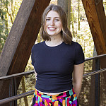 Helena smiling outside on the ravine bridge. She is wearing a black tshirt and floral skirt.