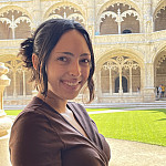 Leah smiling outside a large, white building. She is wearing a brown short-sleeve shirt.