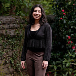 Kaia smiling outside near a wall covered in moss and green bushes with pink flowers.
