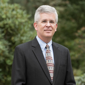 David posing outside. He is wearing glasses, a dark suit with a light shirt, and a red tie.