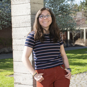 Cloe standing outside on the undergrad campus. She is wearing a blue striped top, red pants, and sandals with socks.