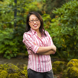 Sharon smiling outside, wearing glasses, a pink and white striped button-down shirt, and gray pants.