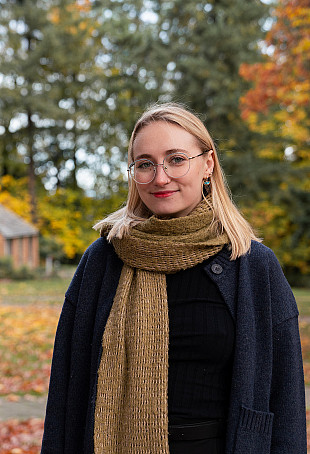 Kindle posing outside the Manor House, wearing a dark coat and brown scarf.
