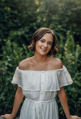 Cass, in a white dress, standing in a forest and smiling.