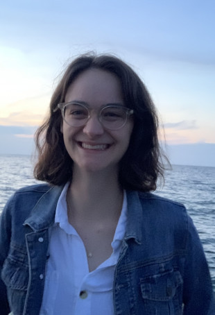Catherine, in a white button down shirt and denim jacket, smiling while standing in front of the ocean at sunset.
