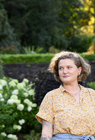 Alicia posing outside in a green garden. They are wearing a yellow top and a blue skirt.
