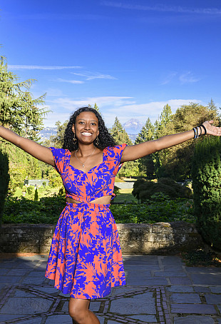 Damaris posing outside with both of her arms raised and Mount Hood in the background. She is wearing a colorful purple and blue dress.