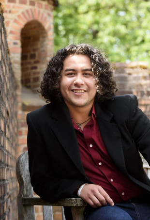 Martin, in a black blazer and red collared shirt, sitting on an outdoor bench next to a brick wall.