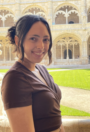 Leah smiling outside a large, white building. She is wearing a brown short-sleeve shirt.