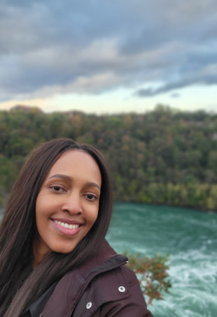 Uwella smiling at the camera, wearing a black jacket, posing in front of a body of water.