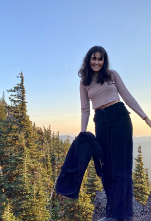 Lilah posing outside, surrounded by trees and a blue sky. She is wearing a light pink long-sleeve top and dark blue jeans.