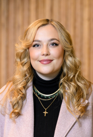 Alie smiling at the camera, wearing a pink blazer over a black turtleneck sweater.