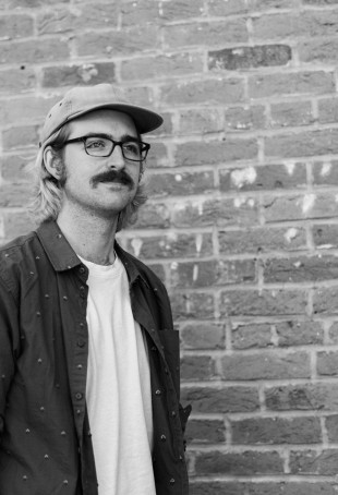 Black and white photo of Jonah. He is wearing a baseball hat, glasses, and a dark button up shirt open over a white tee.