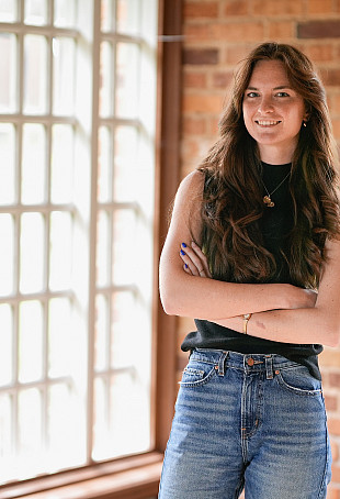 Morgaine posing inside near a window. She is wearing a black tank top and blue jeans.