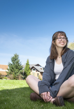 Brynn sitting crosslegged outside on a lawn. She is wearing a shite top, blue cardigan, and denim shorts.