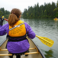 A student in a purple jacket and yellow PFD canoes on a lake with trees in the distance