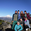 Image shows a group of people posing with a view of Mt Hood and blue skies behind them.