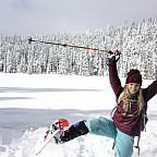Angelina puts her leg in the air while snowshoeing with snow covered trees behind her.
