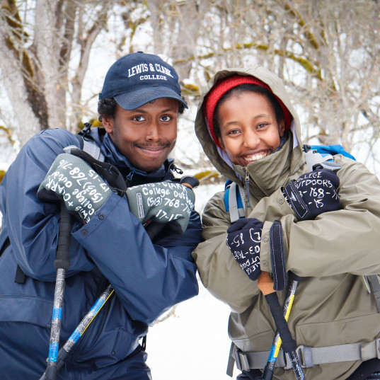 Image Description: Two people are dressed warmly in hats, jackets and mittens, and there is snow behind them. They have their arms crosse...