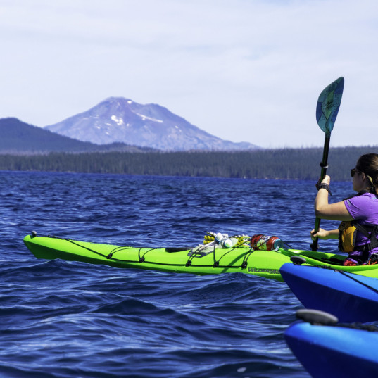 Image Description: Woman in a bright green kayak paddles on a blue lake with mountains in the background