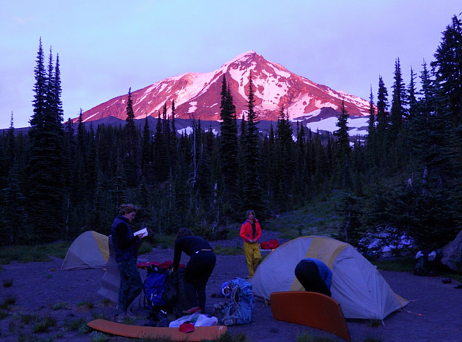 Image shows: a scene of tents and people milling around camp at sunset with Mt Adams lit up pink in the background.