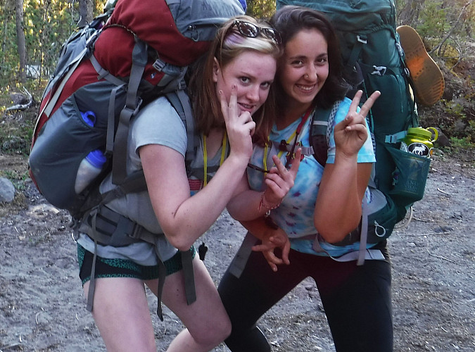 Image shows: two women wearing backpacking packs giving the peace sign and smiling.