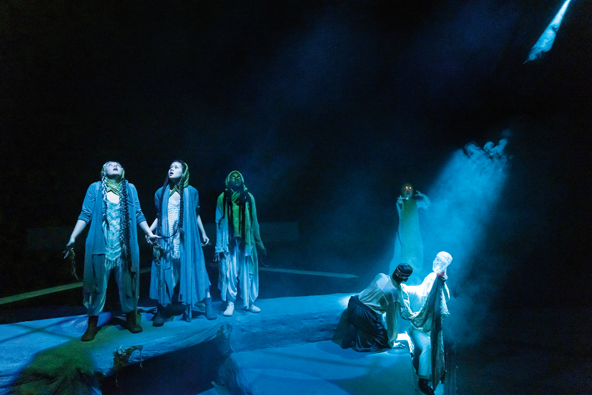 A stage performance, likely a play or a dance piece, is captured. Several individuals are positioned on a flat, bluish-toned surface.