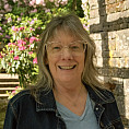 Rebecca is a Caucasian woman with mid length grey hair. She is wear a blue shirt and blue jean jacket. She is smiling under an arbor with...