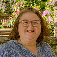 Carolin is smiling and wears glasses. She has short brown hair wearing a blue flower top. In the back ground are pink rhododendrons