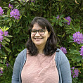 Gaby has mid length brown hair and is wearing a peach shirt with blue cardigan. She is smiling and standing in front of  a purple rhodode...