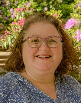 Carolin is smiling and wears glasses. She has short brown hair wearing a blue flower top. In the back ground are pink rhododendrons