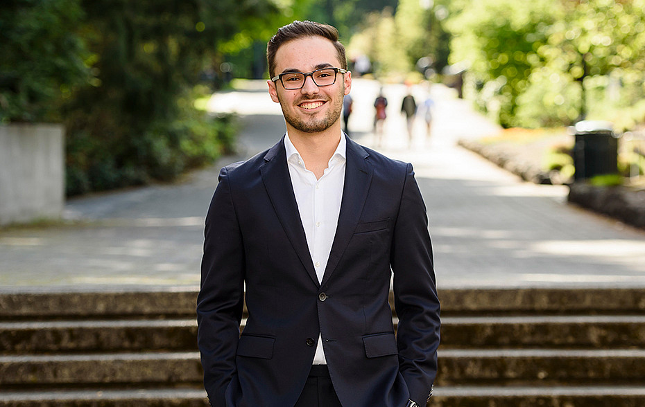 Alex Scioscia BA '21 poses outside wearing a dark suit over a white shirt and no tie.