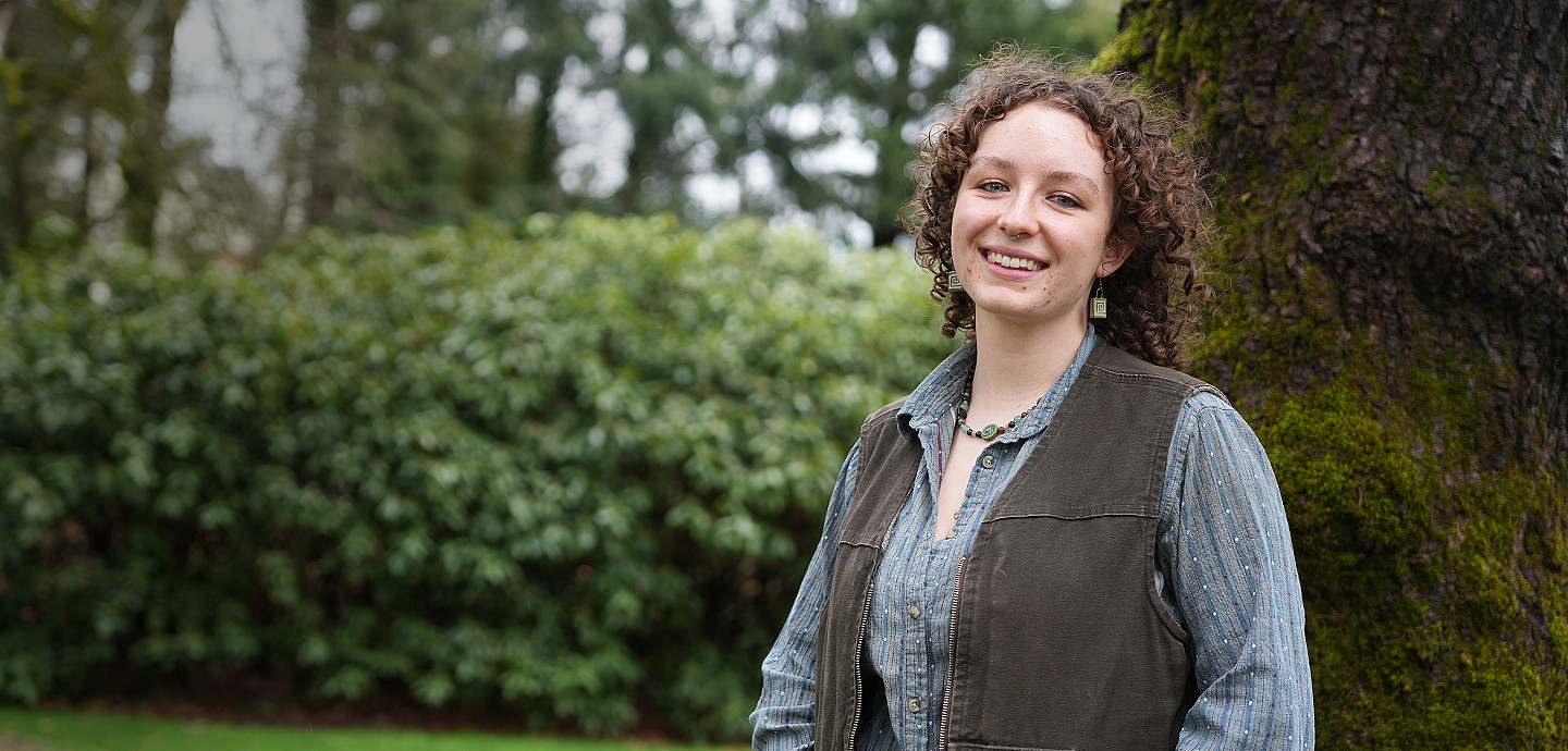 Izz posing outside wearing a brown vest over a button-up shirt.