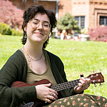 Mack sitting outside Manor House playing a ukulele. They are wearing a green floral dress with a dark green cardigan and black loafers.