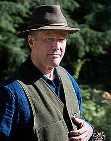 Kim Stafford standing outside, smiling and wearing a brown brimmed hat, green vest, and dark blue shirt.