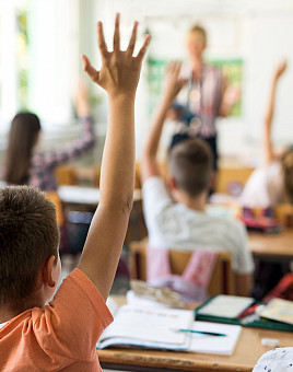 Rear view of little boy and his classmates raising arms to answer teacher's question during the lecture in the classroom.