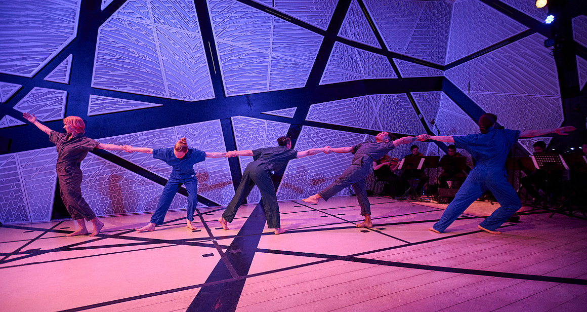 A line of dancers on stage holding hands in front of a colorful background.