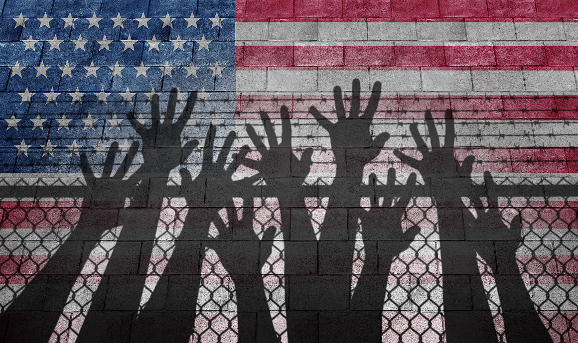 Shadow of hands reaching up over a chain link fence with an American flag as the background.