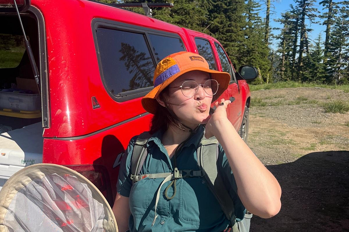 Whitney standing outdoors, wearing a vest and hat, blowing a kiss to the camera.