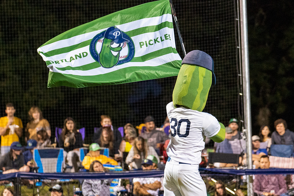 A person in a giant pickle costume wearing a baseball uniform, waving the team flag, while spectators cheer.
