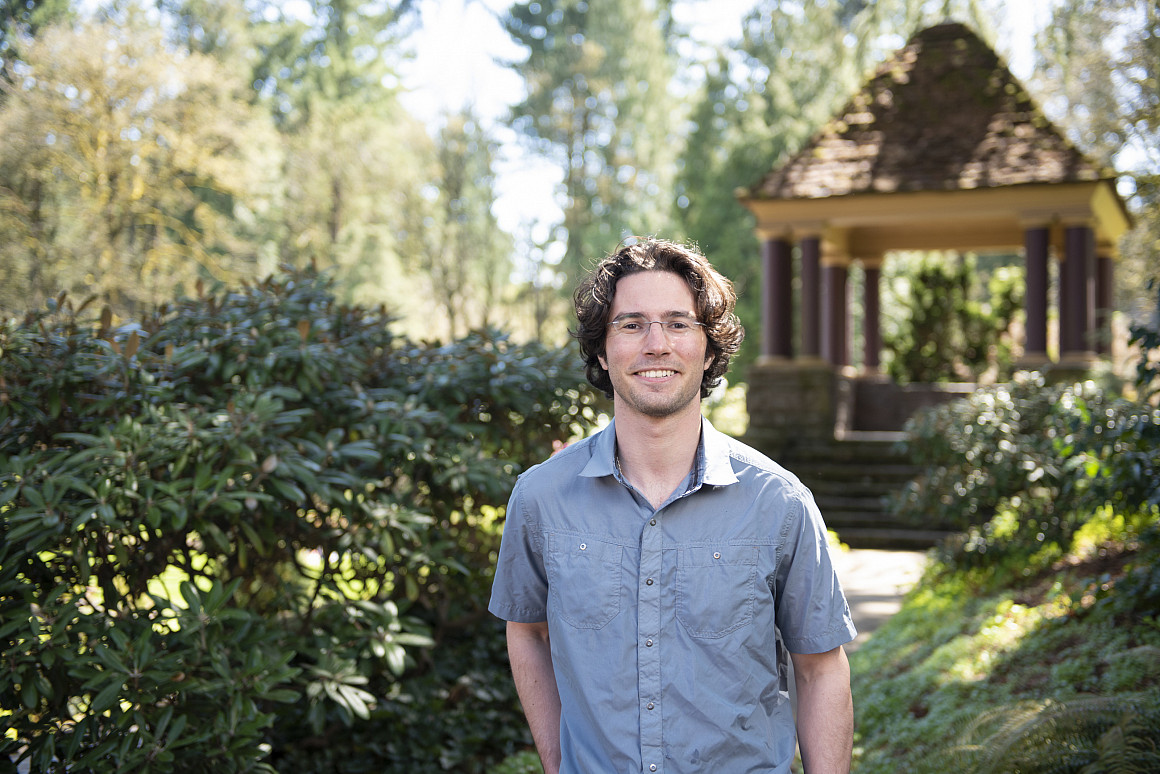 Cooper posing outside near a gazebo, wearing a blue button-up short sleeve shirt and glasses.