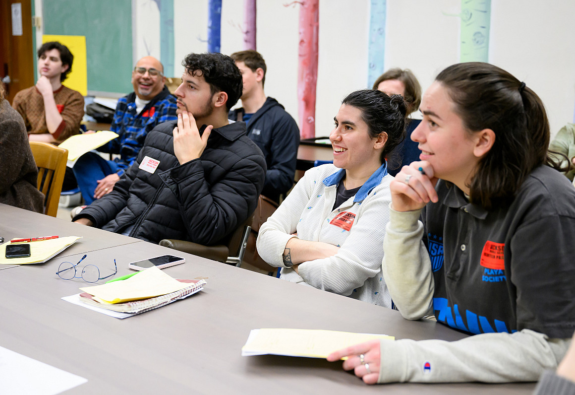 Students sitting together at a table, smiling at the teacher.