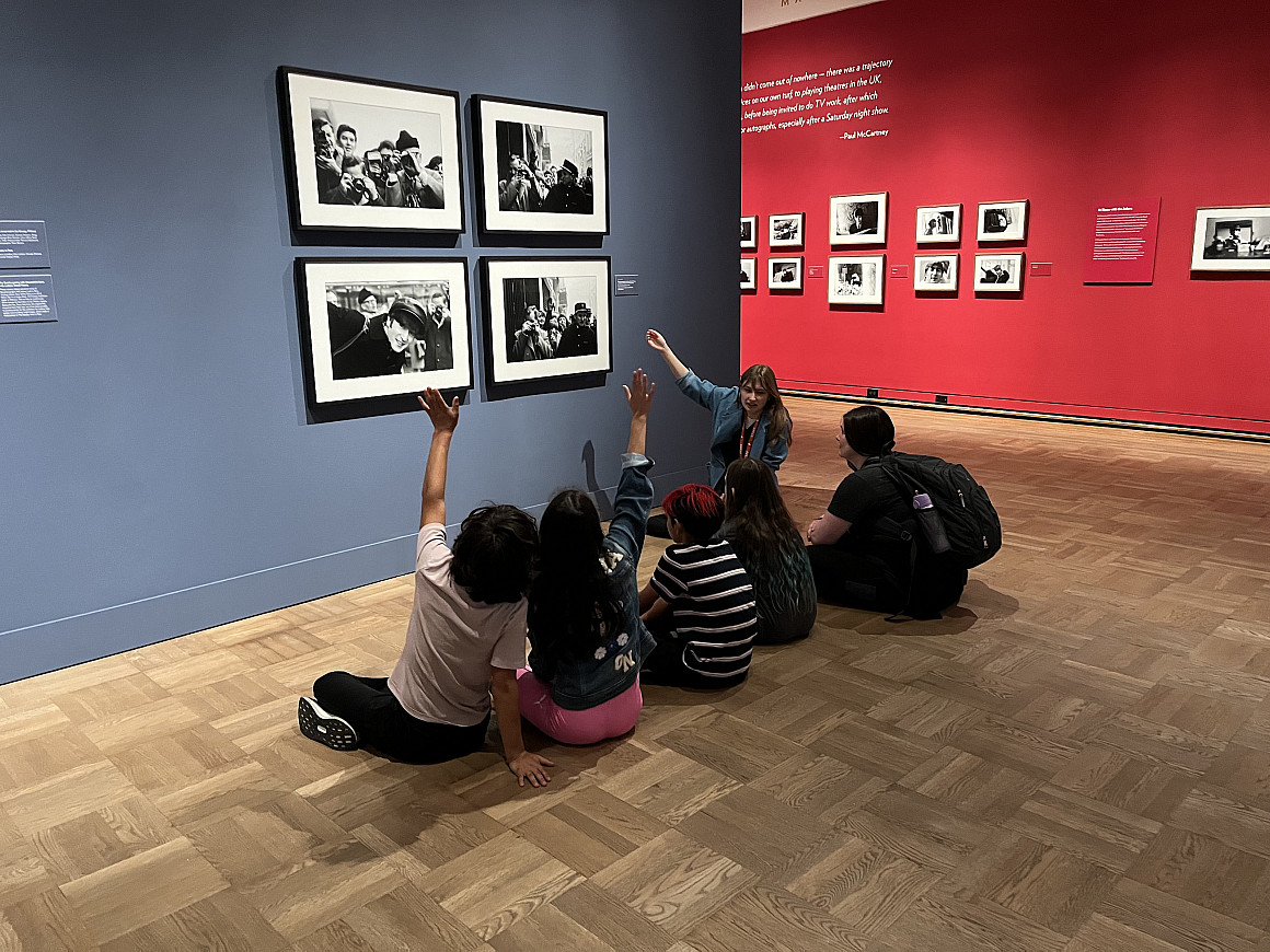 A college student shows elementary children works of art on a wall.