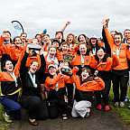 Women's rowing team wearing L&C orange jackets and cheering with their trophy.