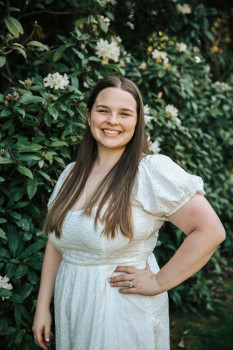 Hannah posing outside. She is wearing a white dress and has one hand on her hip.