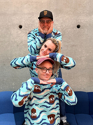 Three people wearing river otter sweaters stacked up on top of each other, smiling.