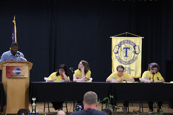 An incarcerated person debating at a lecturn while four students sit at a table.