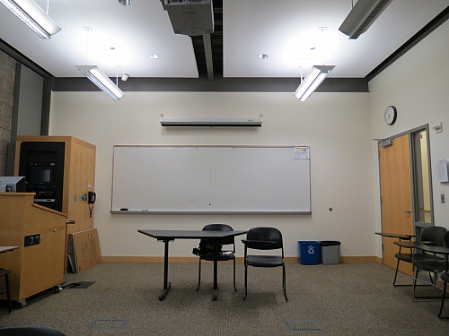 A blank whiteboard is installed on a classroom wall below a rolled-up projection screen. A table and two chairs have been placed in front.